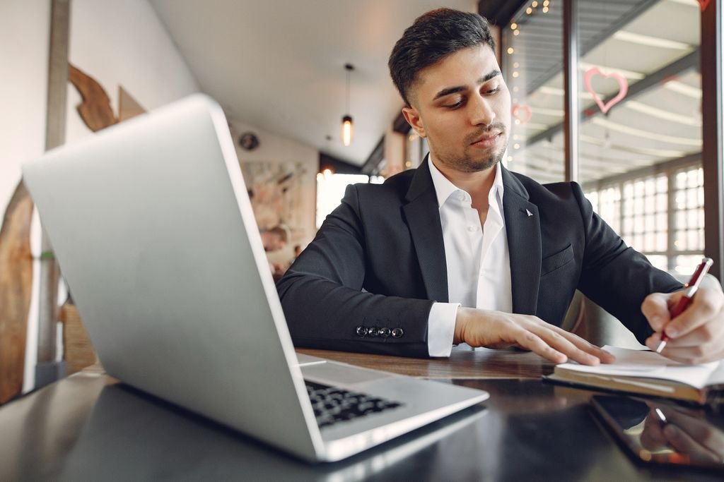 From below of concentrated ethnic male in formal wear sitting in creative workplace and taking notes while using laptop and working on business project